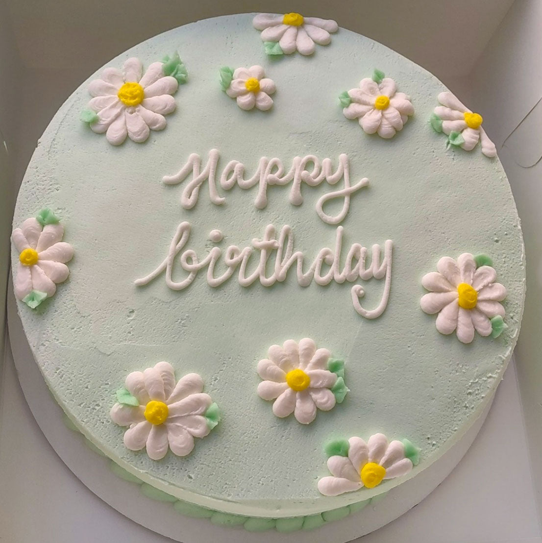 A round mint green icing birthday cake decorated with daisy flowers and 'Happy Birthday' inscribed in the middle.