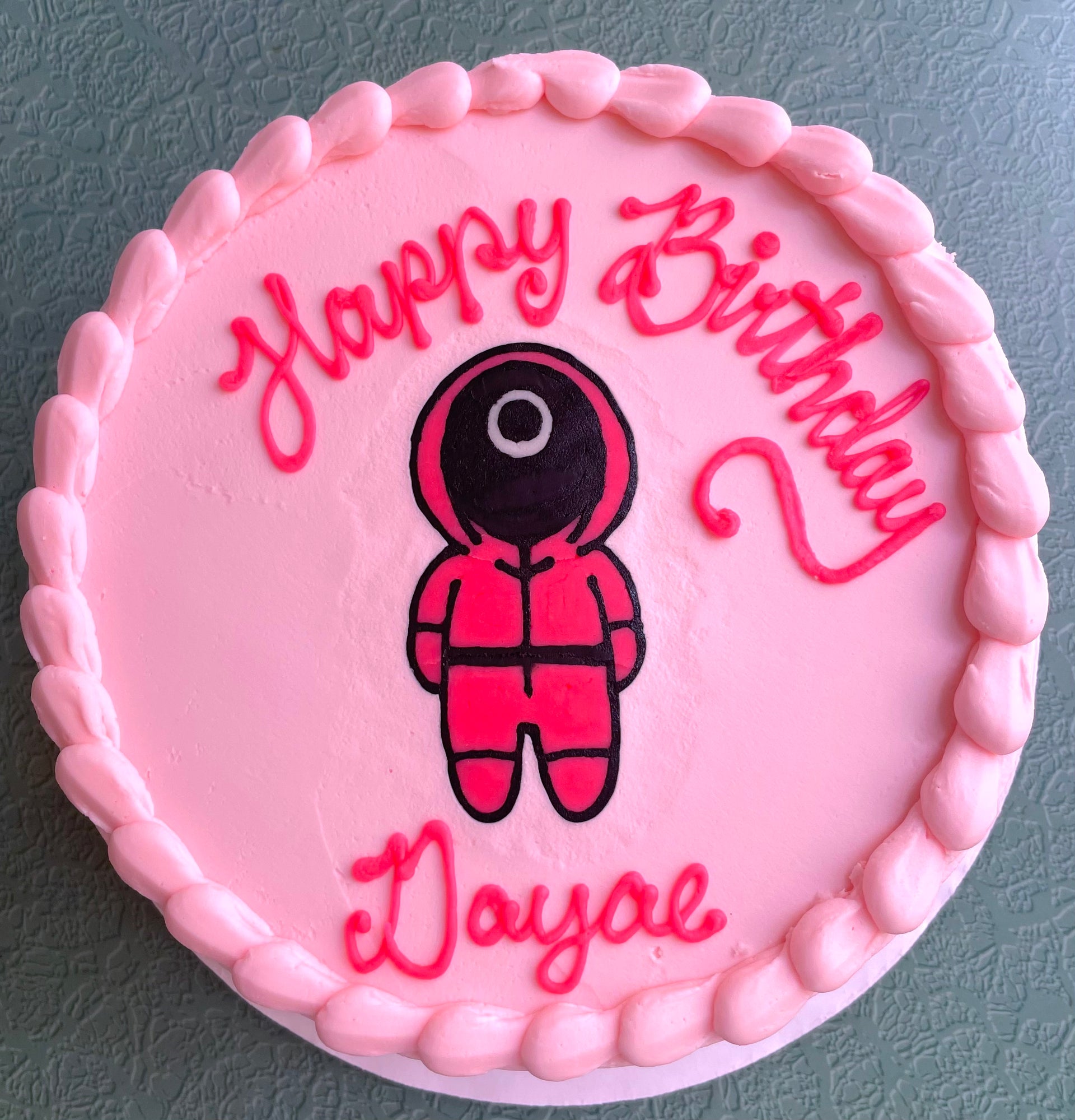 Photo of a round cake frosted in pastel pink buttercream with a hand-drawn image of a Squid Games character. 