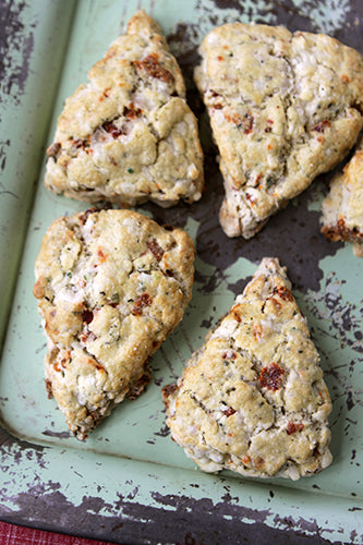 Four sun-dried tomato and Swiss cheese scones on a green surface, with one scone cut in half to show the filling.