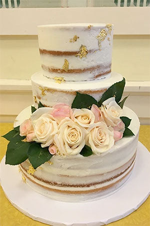 A 3-tier wedding cake frosted in buttercream with a semi-naked finish. Adorned with customer-supplied fresh roses and greenery. Gold leaf is splattered around the sides of the cake. 