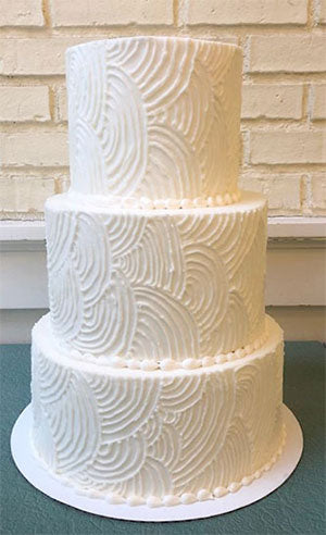 Three-tiered white wedding cake with textured buttercream frosting featuring a wavy, swirled pattern.