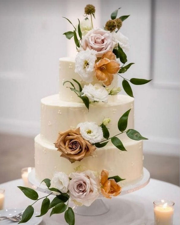 Three-tiered white wedding cake adorned with blush pink, dark orange, and white roses, and green foliage, on a white cake stand with a white candle in the right corner.