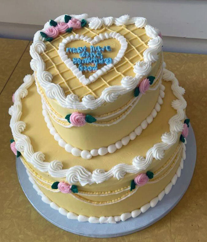 A two-tiered heart-shaped cake with yellow frosting and white piped details, adorned with pink roses and green leaves, featuring a lattice pattern on the top tier and pearl-like borders, with a blue inscription "Must have done something good" on the top heart.