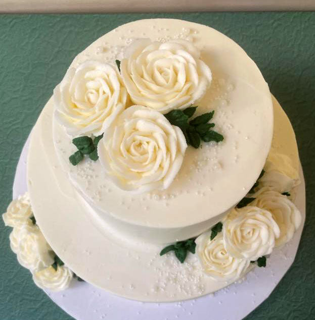 Two-tiered white buttercream cake decorated with numerous ivory roses and green leaves on a blue textured surface.