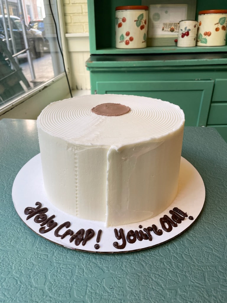 A custom decorated birthday cake with white buttercream frosting, placed on a plate with a written message around the base, displayed on a green surface.