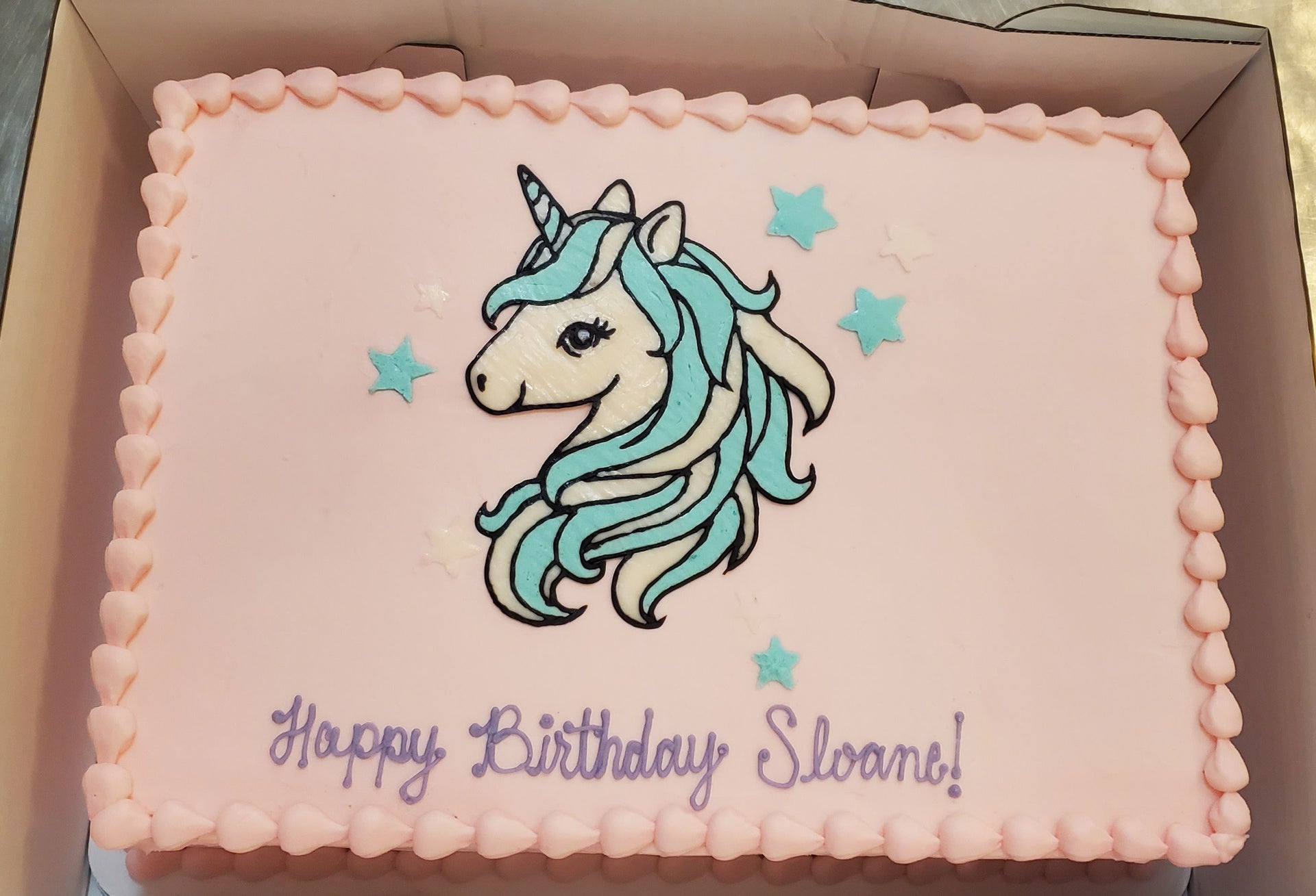 A rectangular pink birthday cake with a detailed unicorn head illustration in blue and white, surrounded by light blue stars, and the text "Happy Birthday Sloane!" written in purple script at the bottom.