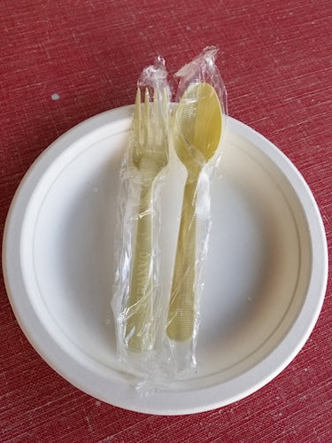 Disposable white plate with a wrapped set of a biodegradable fork and spoon, light yellow in color, on a red surface.