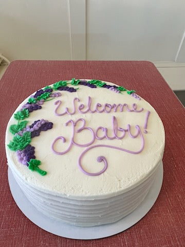 Round cake with 'Welcome Baby!' text and green and purple decorations on a red surface.