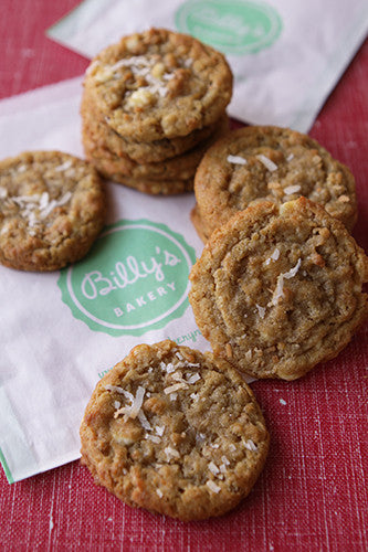 White chocolate and coconut cookies from Billy's Bakery, with a stack of cookies on a white paper bag featuring the 'Billy's Bakery' logo, all resting on a red textured surface.