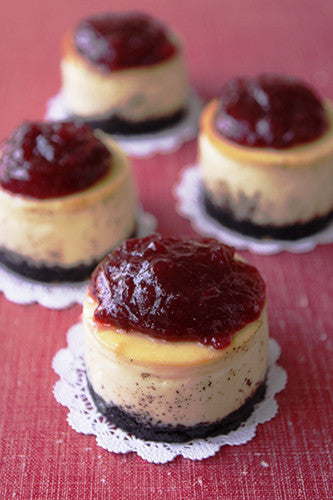 Four mini cheesecakes with dark cookie crusts and a generous topping of red berry compote, arranged on white lace doilies on a red textured surface. 
