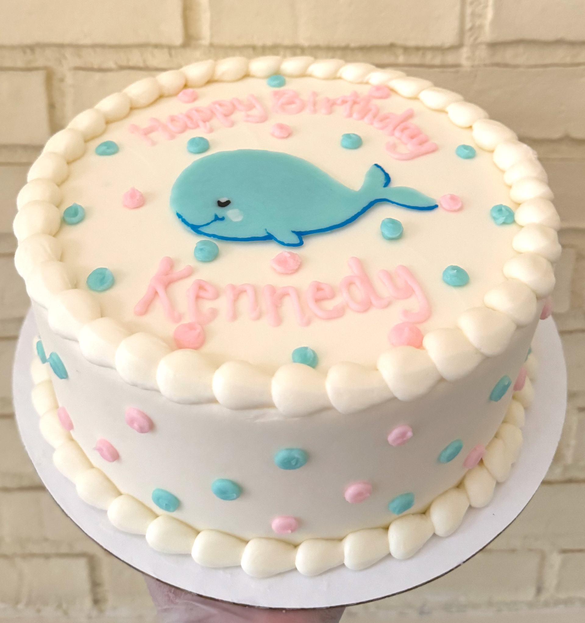 White birthday cake with a blue whale decoration, pink and blue sprinkles, and 'Happy Birthday Kennedy' written in pink frosting.