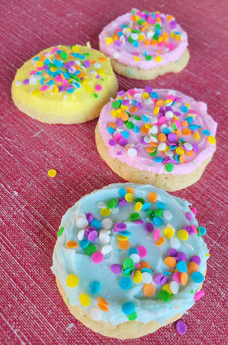 A photo showing three cookies with colorful sprinkles on top, frosted with different colored buttercream frosting. 