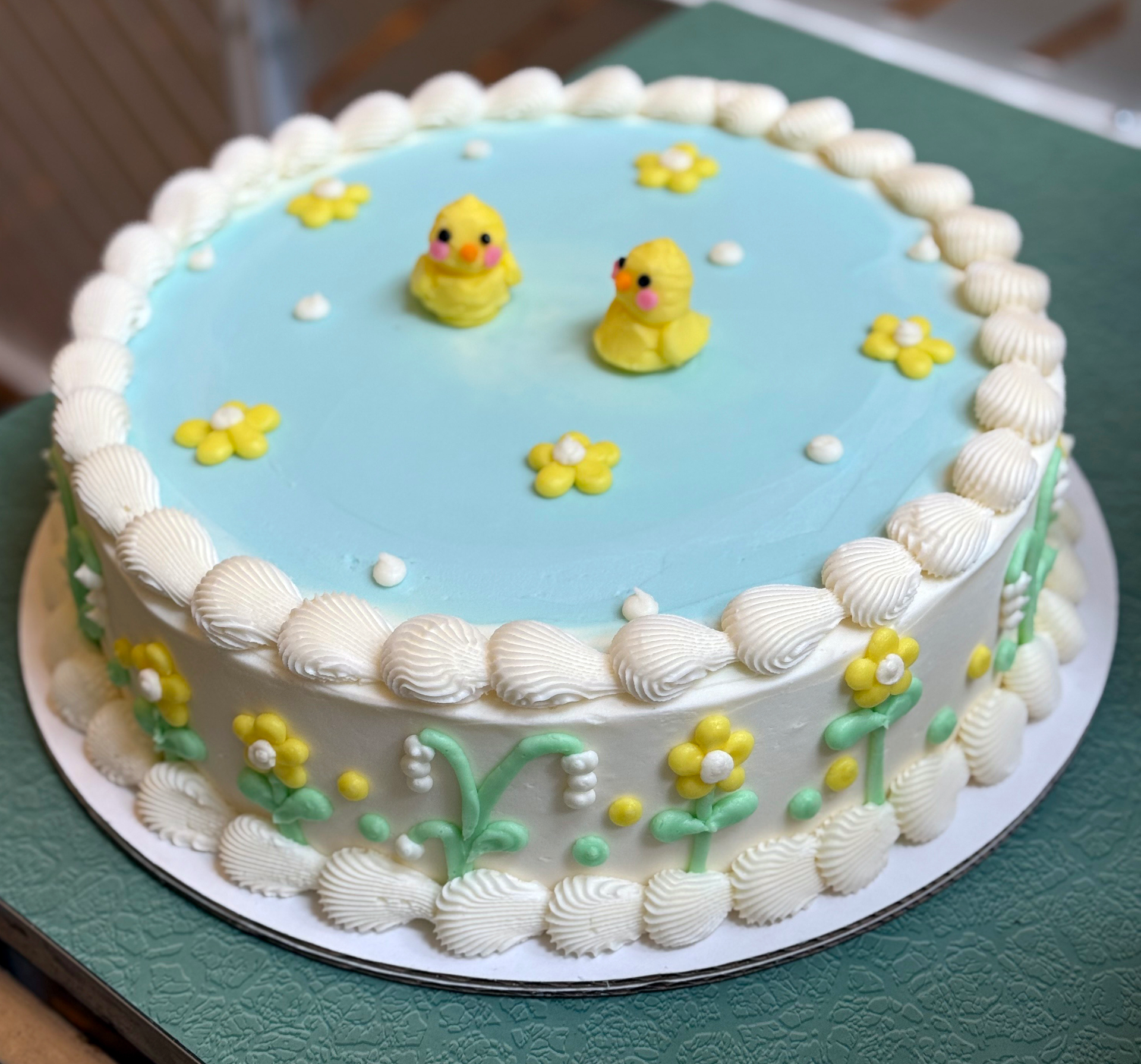 A decorated cake with white buttercream frosting and blue buttercream, adorned with yellow daisy-like flowers and small sugar ducks.