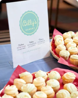 A sign for Billy's Bakery with locations and contact information alongside trays of cupcakes.