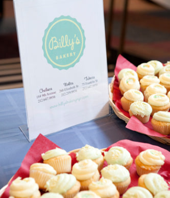 A menu board from Billy's Bakery advertising cupcakes and other baked goods, positioned next to a basket filled with small, frosted cupcakes.