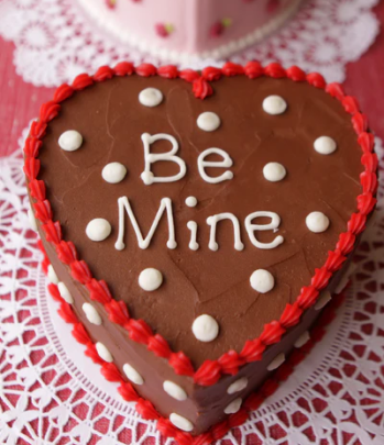 Heart-shaped chocolate cake with 'Be Mine' written in white icing, decorated with white polka dots and a red piped border.