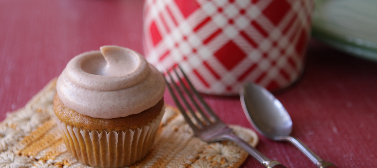 Pumpkin spice cupcake with cinnamon cream cheese frosting on a crocheted placemat next to a fork, spoon, and red and white patterned mug.