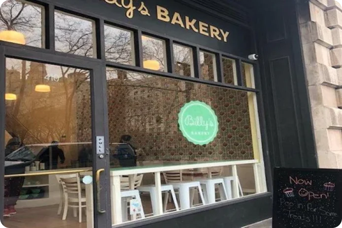 The exterior storefront of Billy's Bakery, featuring a glass door with the street number 410, large windows, and a round green "Billy's BAKERY" logo visible inside.