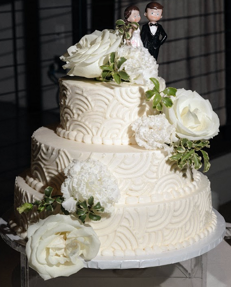 Three-tiered white wedding cake with white roses and swirls, greenery, and a bride and groom topper.
