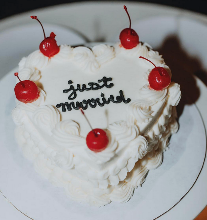 A heart-shaped white cake with "just married" written in black icing, topped with red maraschino cherries.