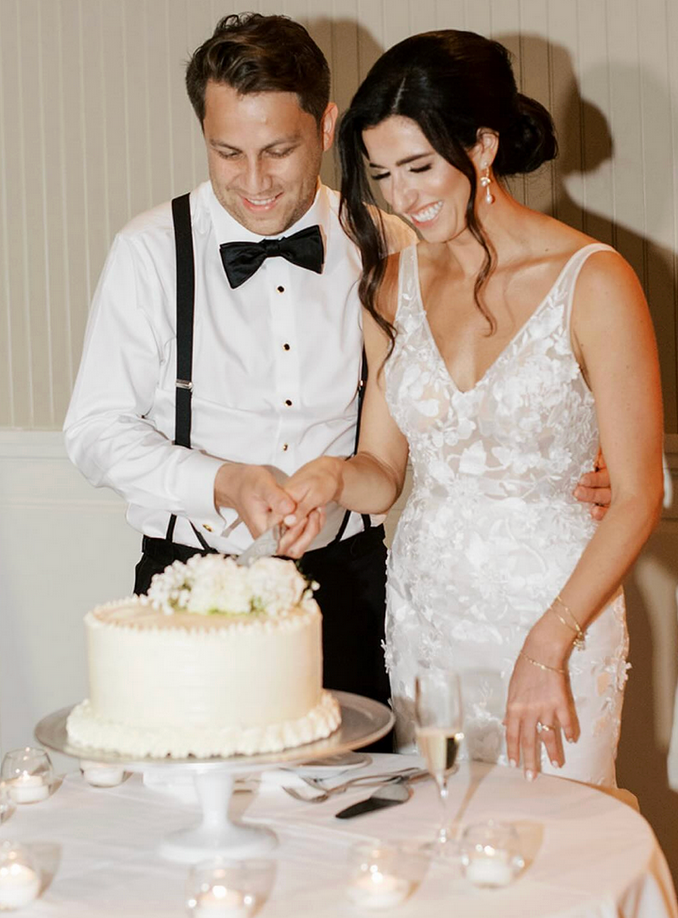 A smiling bride and groom in wedding attire cut a white wedding cake adorned with white flowers at their reception.
