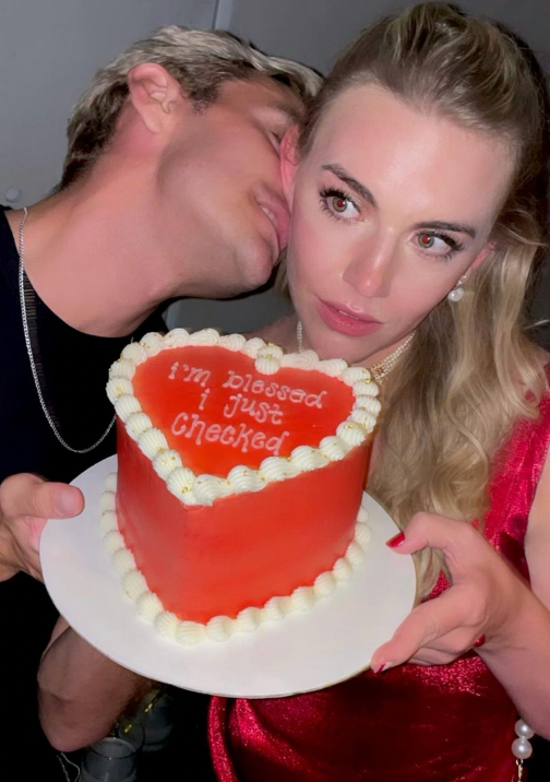 A heart-shaped cake with the phrase 'I'm Blessed I Just Checked' written on it, being held by a person with a blurred face and a woman looking at the camera.