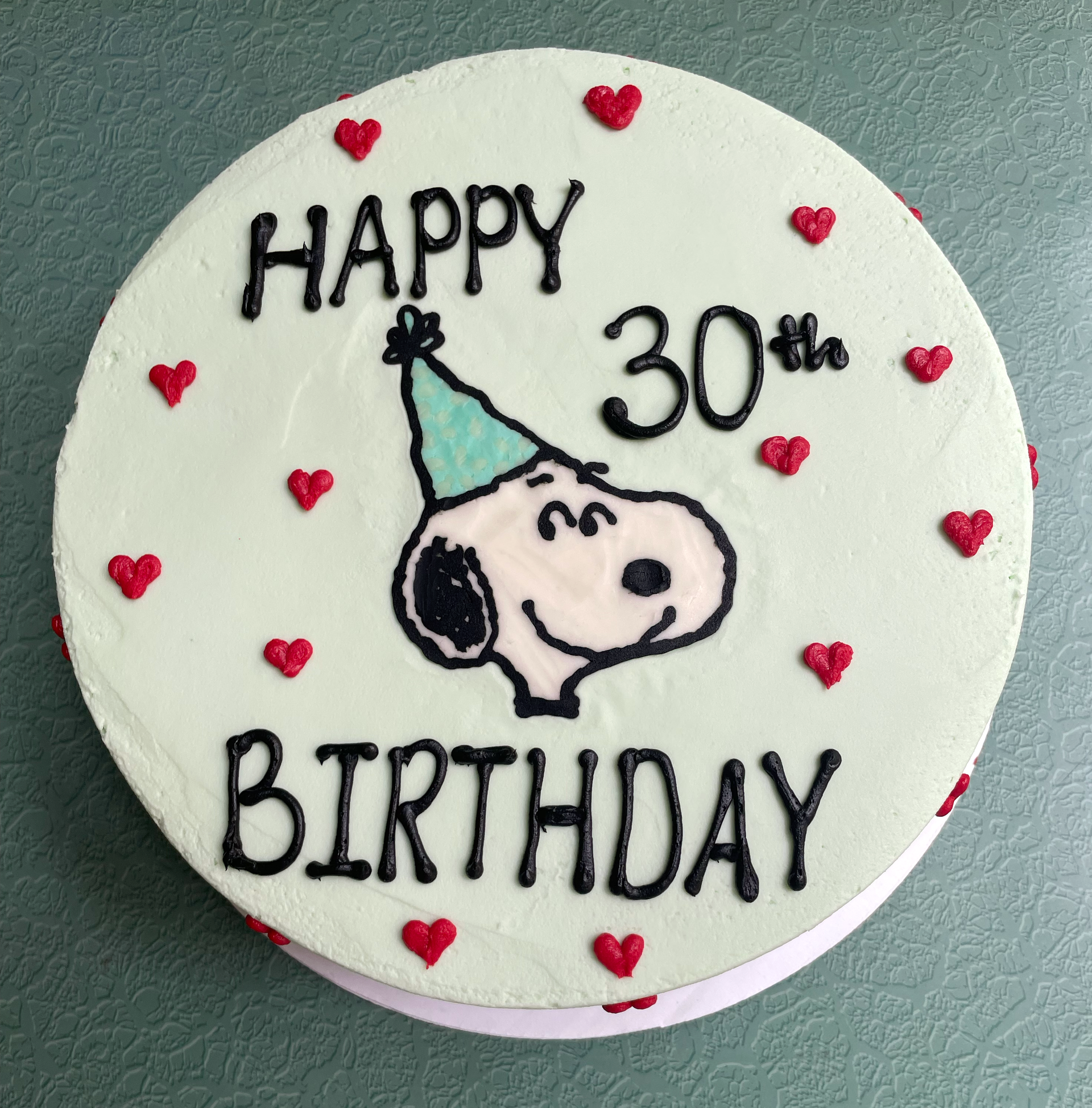 A round, pastel green frosted birthday cake features a hand-piped image of Snoopy from the Peanuts cartoon, wearing a blue party hat. The text "HAPPY 30th BIRTHDAY" is written in bold black icing above and below Snoopy. The cake is decorated with small red buttercream hearts evenly spaced around the top and sides, giving it a festive and whimsical appearance. The cake sits on a textured green background, enhancing its cheerful design. Perfect for a 30th birthday celebration with a nostalgic, cartoon-themed 