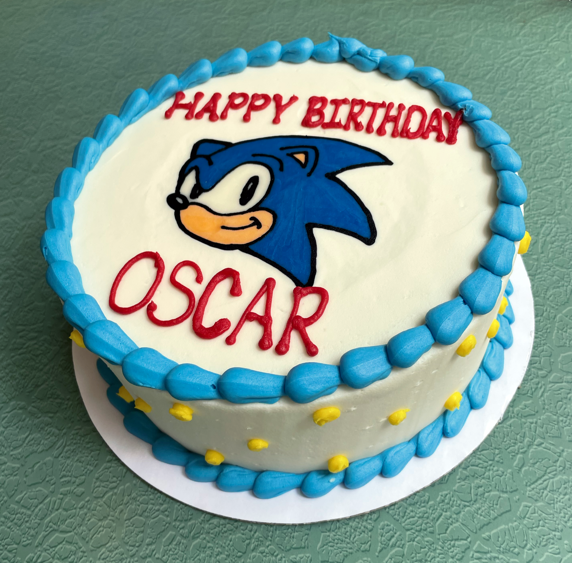 A decorated cake with white buttercream frosting adorned with a hand-drawn Sonic the Hedgehog character. Yellow polka dots on the sides of the cake. 