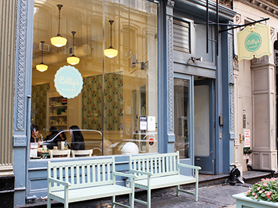 Exterior of Billy's Bakery in Tribeca, NYC, showing the storefront with two light green benches outside and a view of the interior with pendant lights and customers.